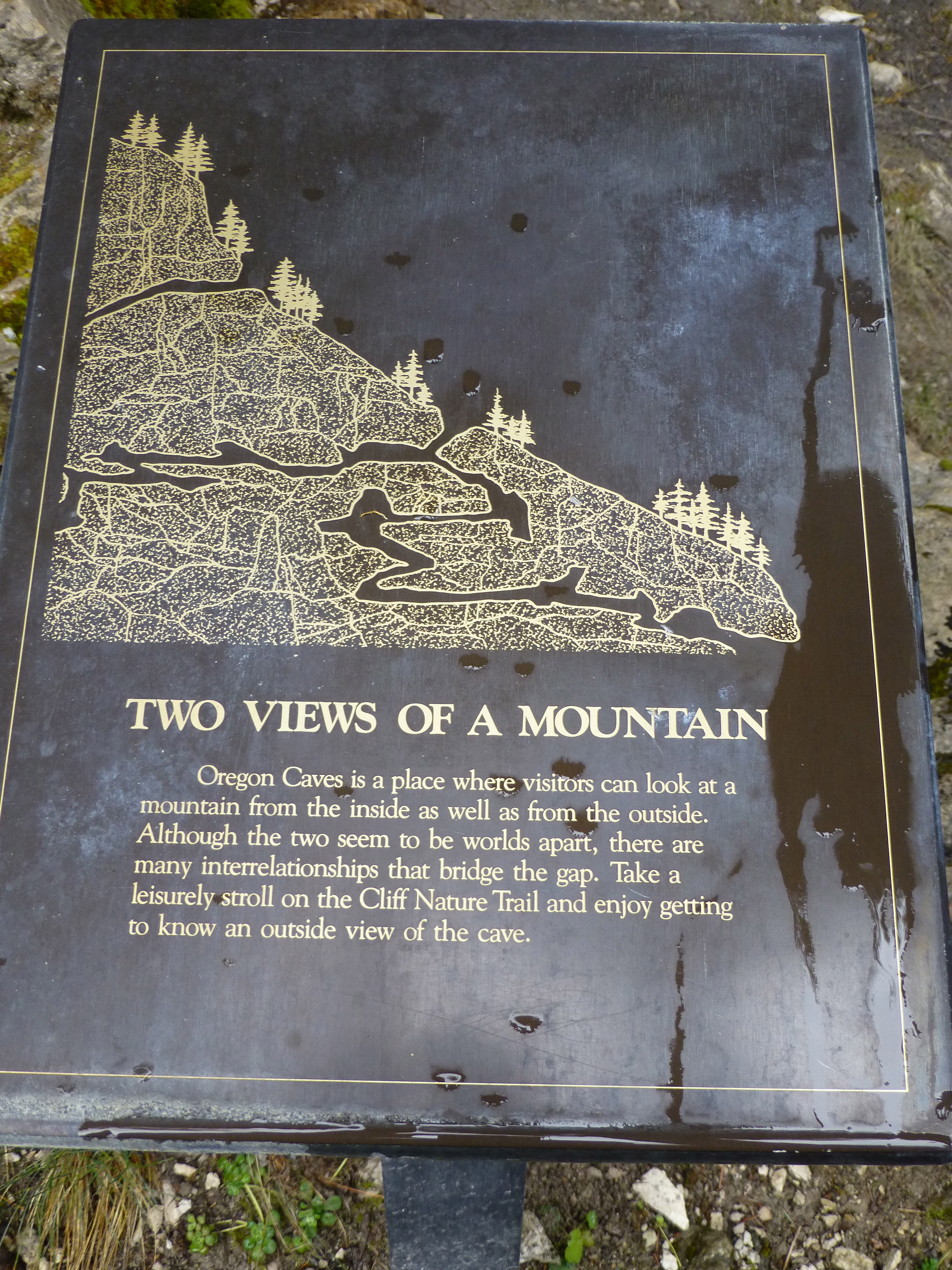 A sign titled 'Two Views of a Mountain' describing Oregon Caves, featuring a gold embossed illustration of a mountain landscape with trees and rocks.