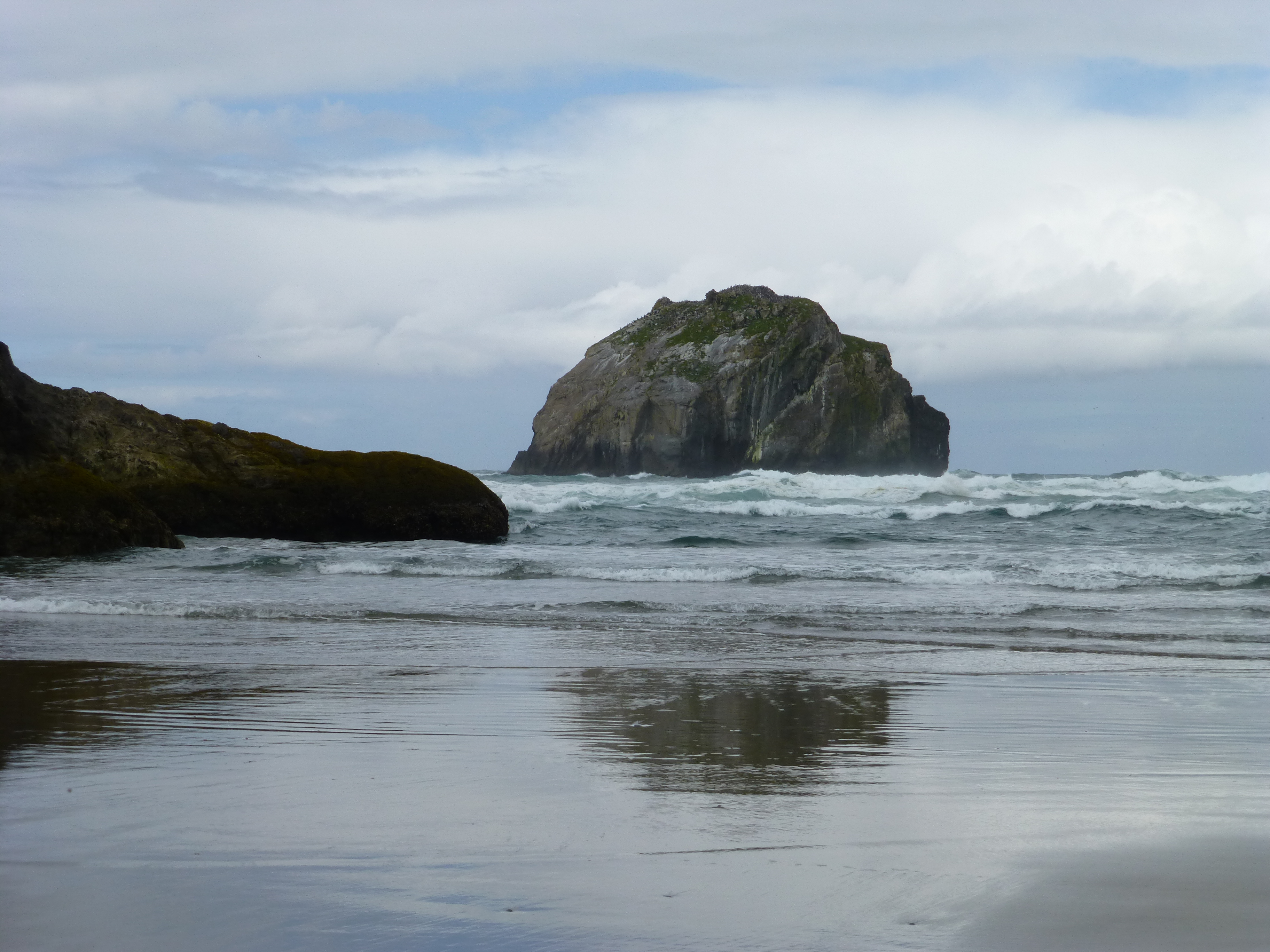 A rocky coastline with a large sea stack in the distance, waves crashing onto the shore under a cloudy sky.