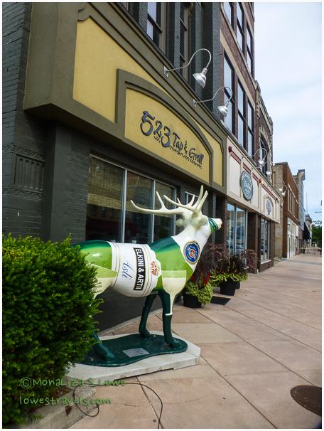 A colorful, painted deer statue wearing a beer bottle as a body stands in front of a restaurant with various storefronts in the background.