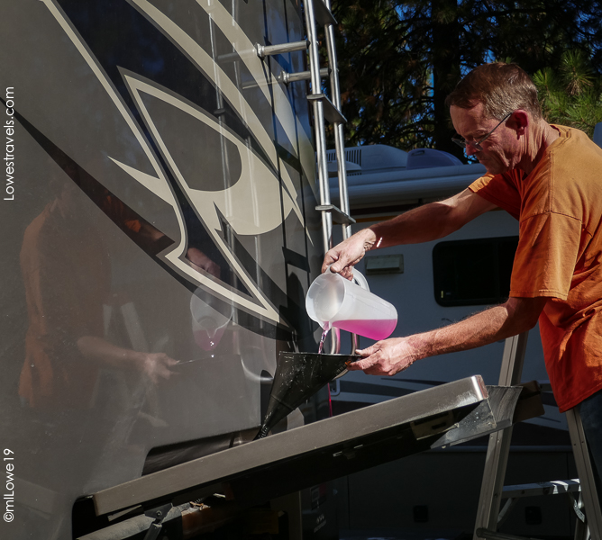 A person pours a pink liquid from a container into a funnel while working on the side of an RV.