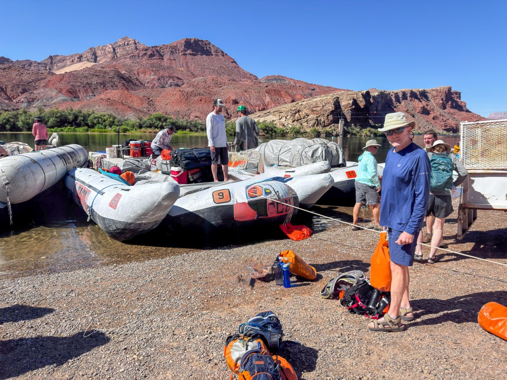 People preparing for a motorized rafting trip on the Colorado River, with rafts and gear at the launch site against a backdrop of red canyon cliffs.