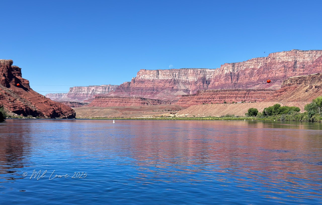 A scenic view of the Colorado River at the Grand Canyon, showcasing the majestic red rock formations and clear blue skies, with reflections in the water.
