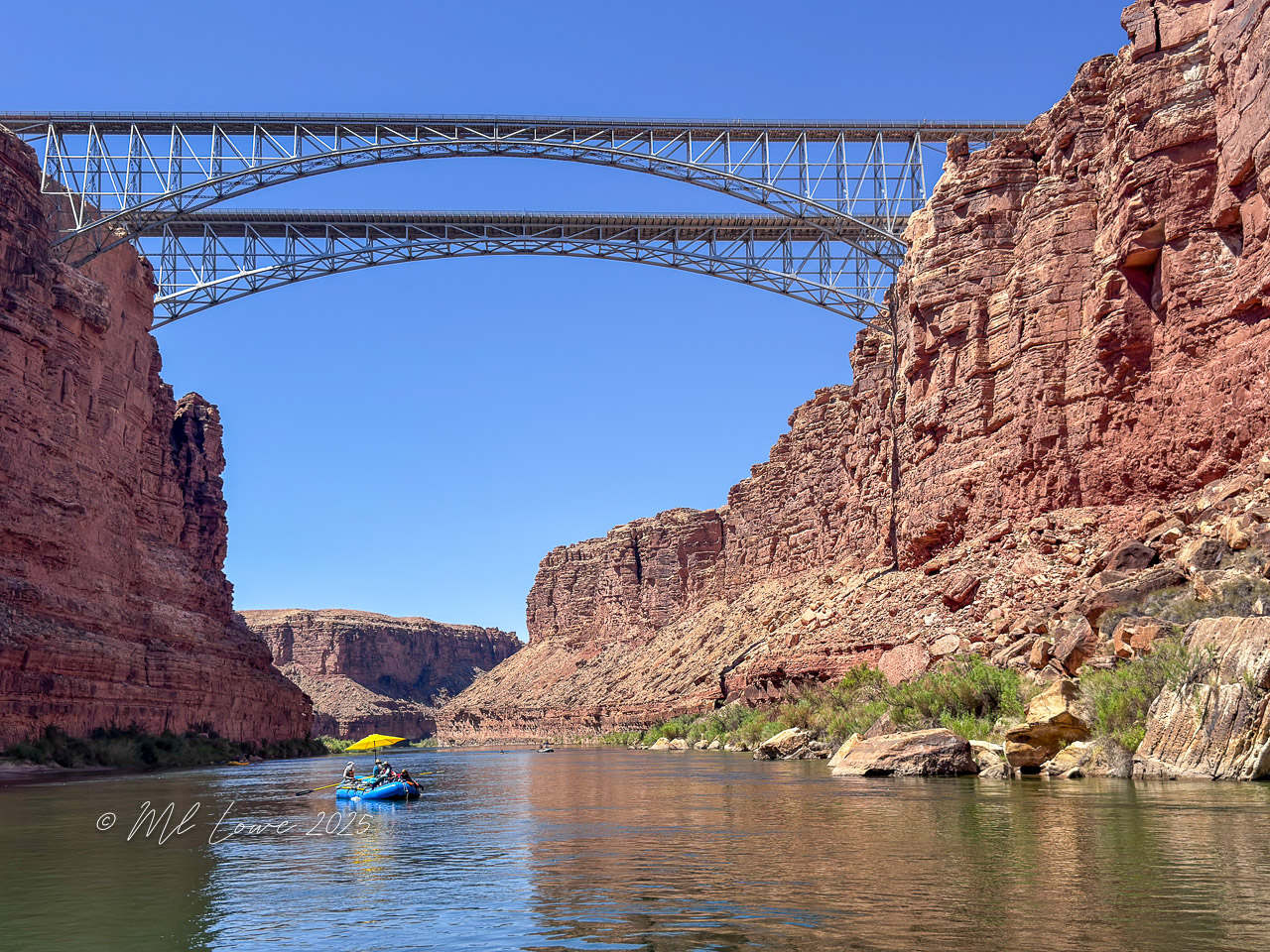 A view of a blue raft on the Colorado River at the Grand Canyon, with red rock cliffs and a metal bridge overhead against a clear blue sky.
