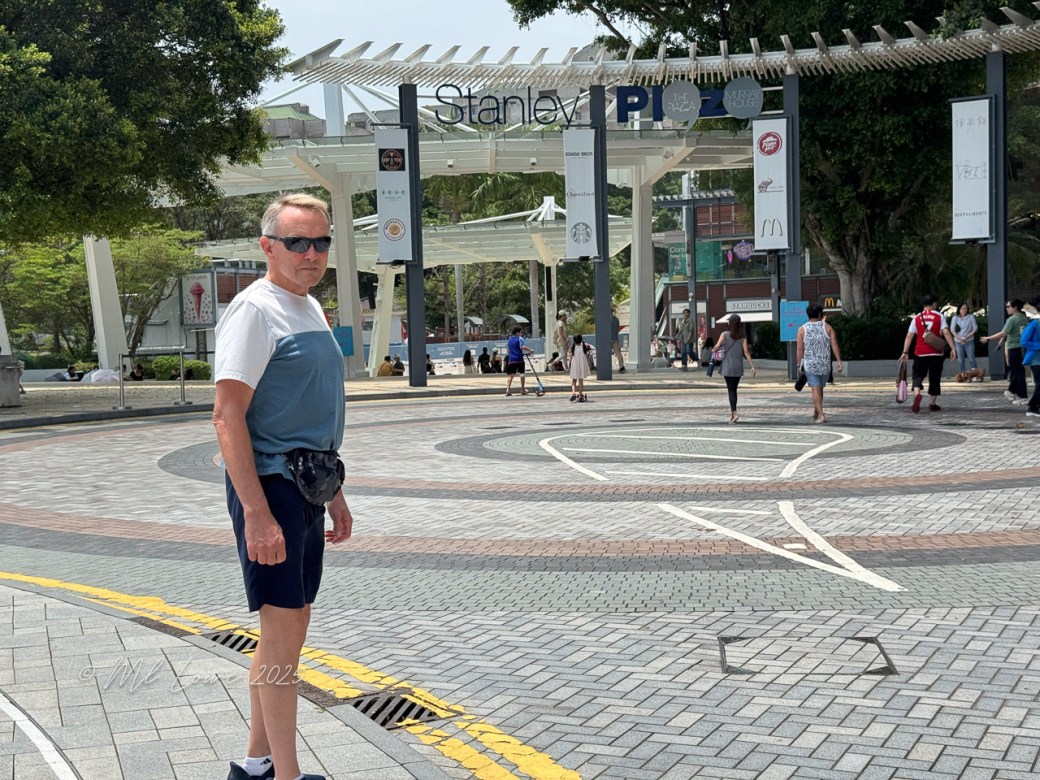 A man stands in front of the entrance to Stanley Plaza in Hong Kong, wearing sunglasses and a casual outfit. The area is bustling with people walking around the circular plaza, with shops visible in the background.