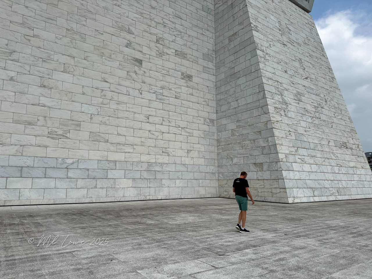 A person standing on a stone plaza, under a large, modern architectural structure with a textured marble wall.