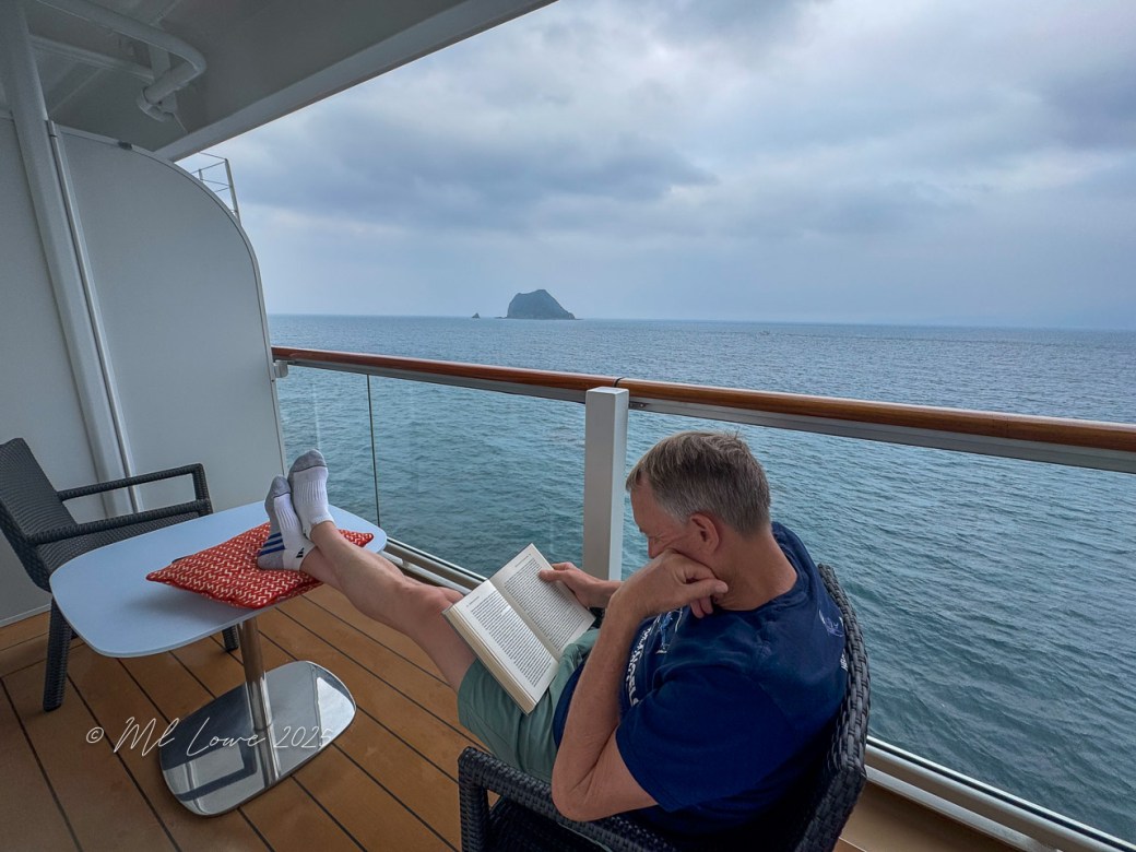 A person reading a book while relaxing on a cruise ship balcony with ocean views in the background.