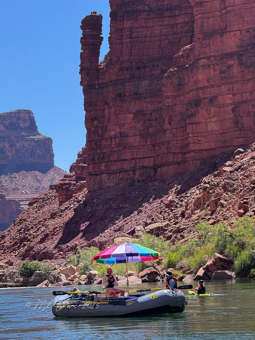 A motorized raft with a colorful umbrella on the Colorado River, with towering red rock formations in the background.