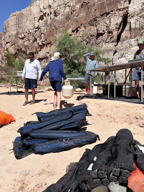 Group of people preparing camping gear along a sandy shore near canyon walls, with one person carrying a container and others standing nearby.