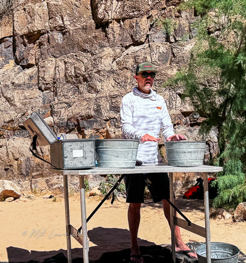 A man standing at a table outdoors, set up with metal wash basins, in front of a rocky cliff backdrop.