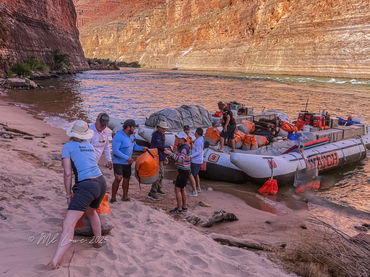 Group of people unloading gear from a motorized raft on the sandy banks of the Colorado River at the Grand Canyon, surrounded by towering canyon walls.