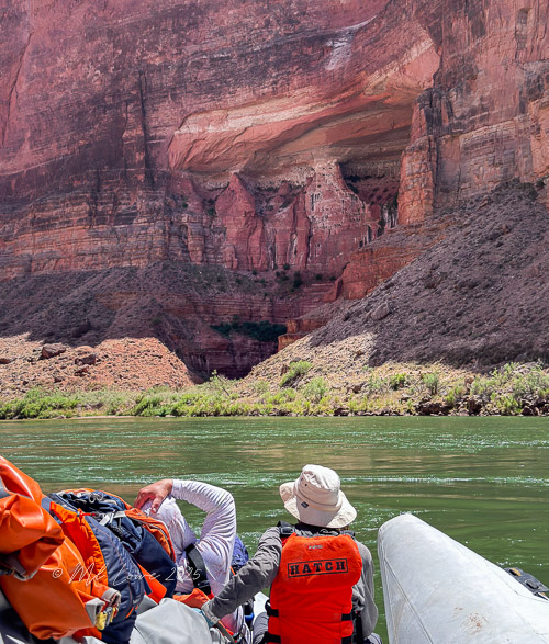 Two adventurers on a motorized raft on the Colorado River, exploring the Grand Canyon, with dramatic red rock formations in the background.