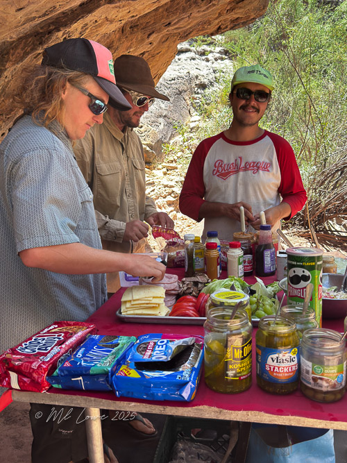 Three men preparing food on a river rafting trip, surrounded by various ingredients and snacks on a table set under a rocky overhang.