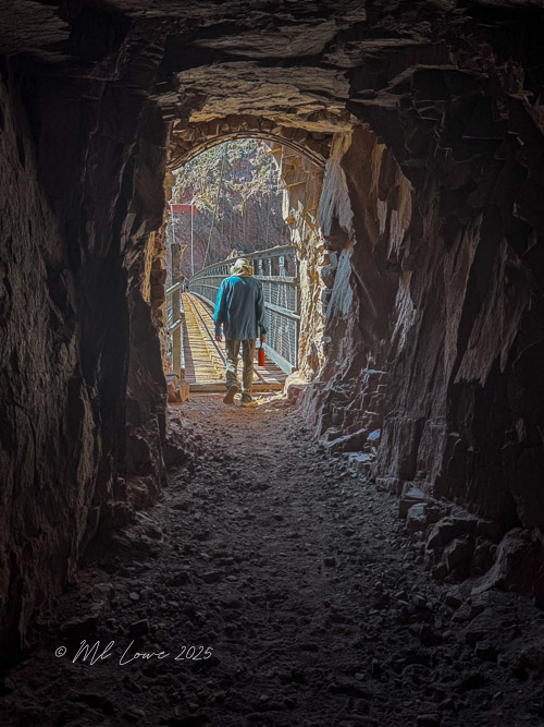 A person walking through a dark tunnel with rocky walls, leading to a bright opening that reveals a path with railroad tracks.