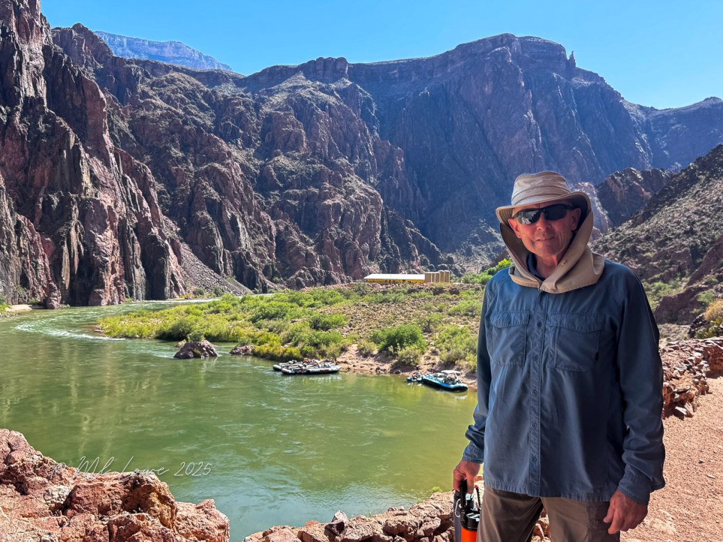 A person standing near the Colorado River in the Grand Canyon, with towering rock formations in the background and rafts visible in the water.