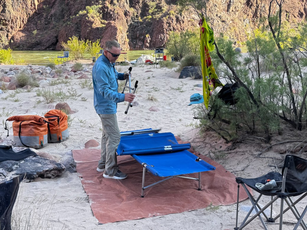 A person setting up a blue cot on a sandy riverbank with bags nearby, surrounded by greenery and rocky cliffs in the background.