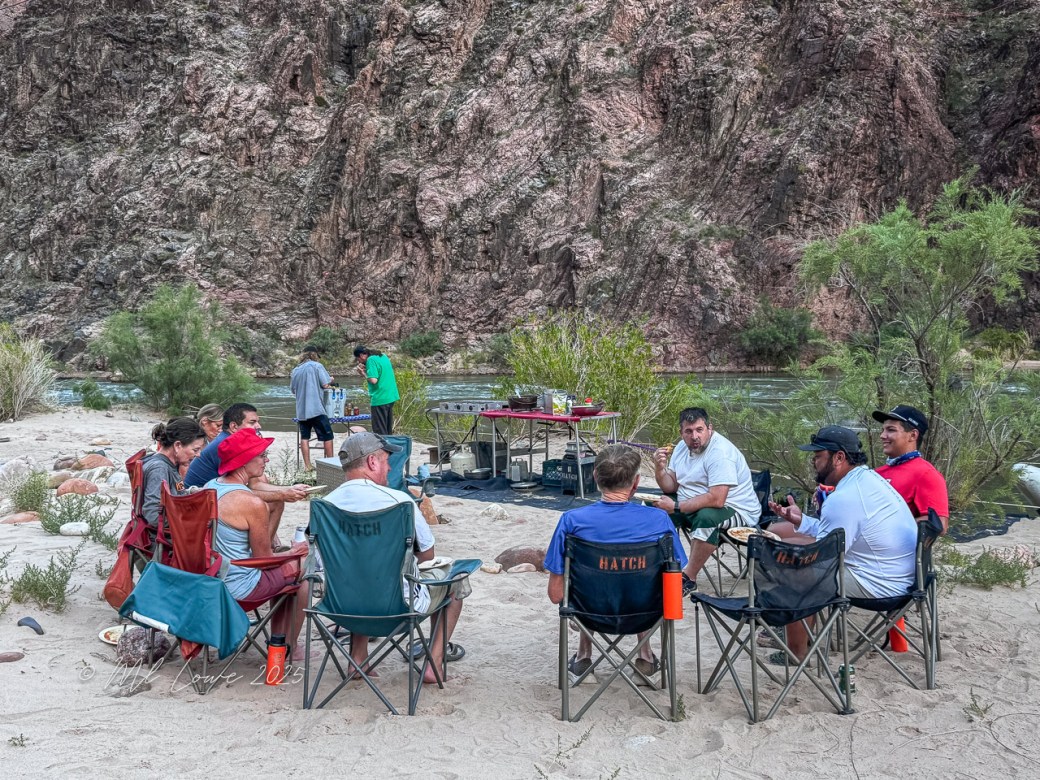 Group of people sitting around a campfire in folding chairs by the Colorado River, enjoying a meal while surrounded by scenic canyon walls.