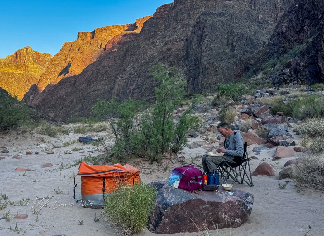 A person sitting on a camp chair, enjoying a meal, surrounded by boulders and vegetation in a canyon landscape with sunlit cliffs in the background.