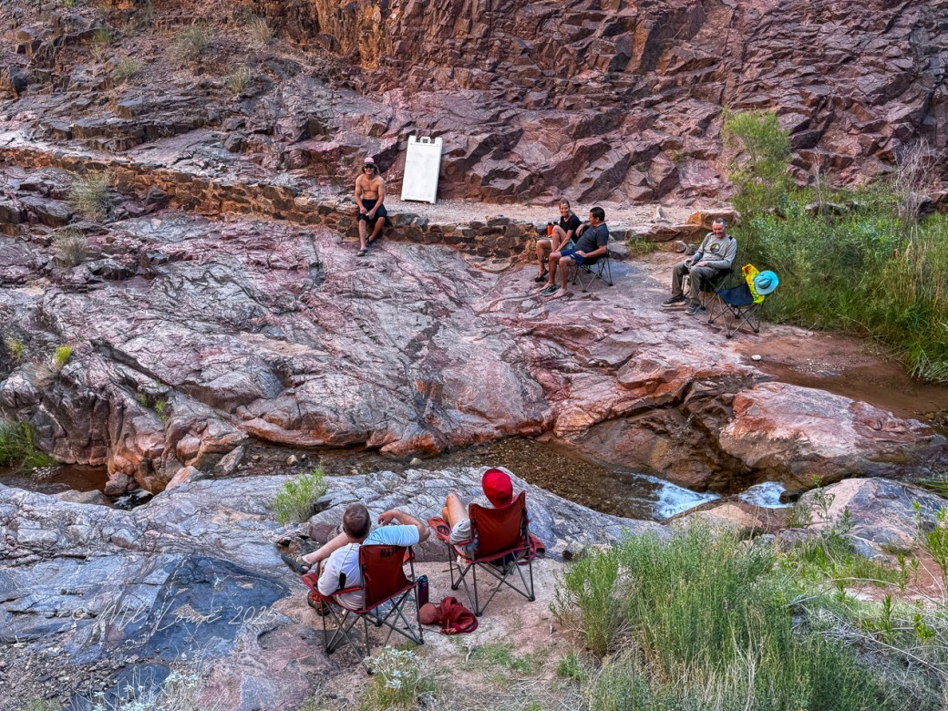 A group of people relaxing by a stream in the Grand Canyon, sitting on camping chairs surrounded by red rock formations and greenery.