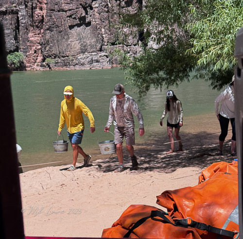 Three individuals walking on a sandy riverbank, carrying buckets and wearing sun protection clothing, with a river and rocky cliffs in the background.