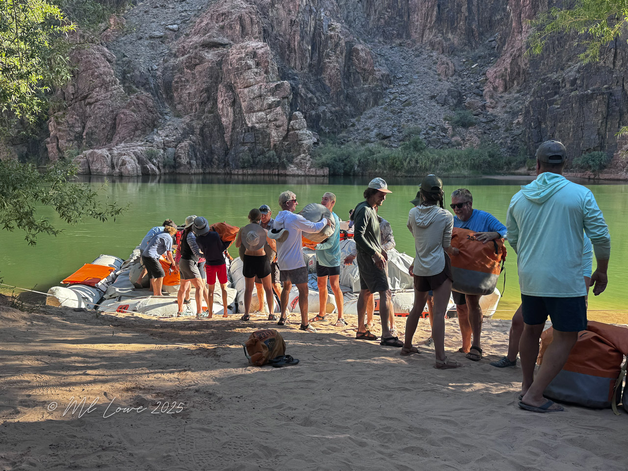 Group of people preparing gear on the sandy riverbank beside the water, with rocky canyon walls in the background.