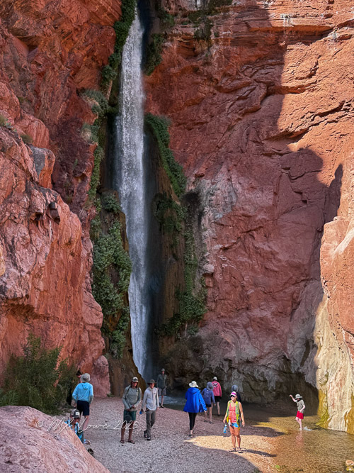 Hikers exploring a rocky canyon with a waterfall in the background, surrounded by red rock formations.