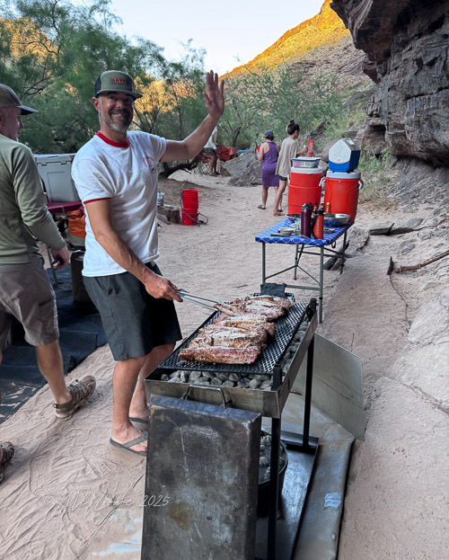 A man grilling meat outdoors in a canyon setting, smiling and waving, with camping gear and red containers in the background.