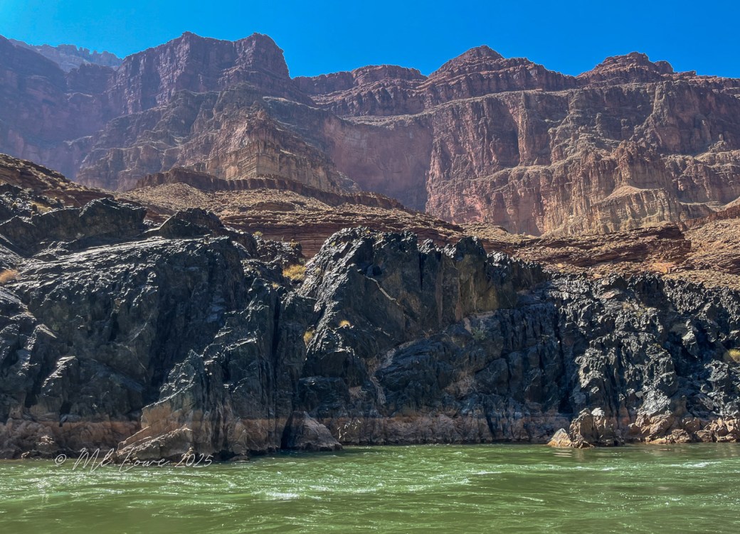 A view of the Colorado River with towering red rock cliffs of the Grand Canyon in the background, reflecting the bright blue sky above.