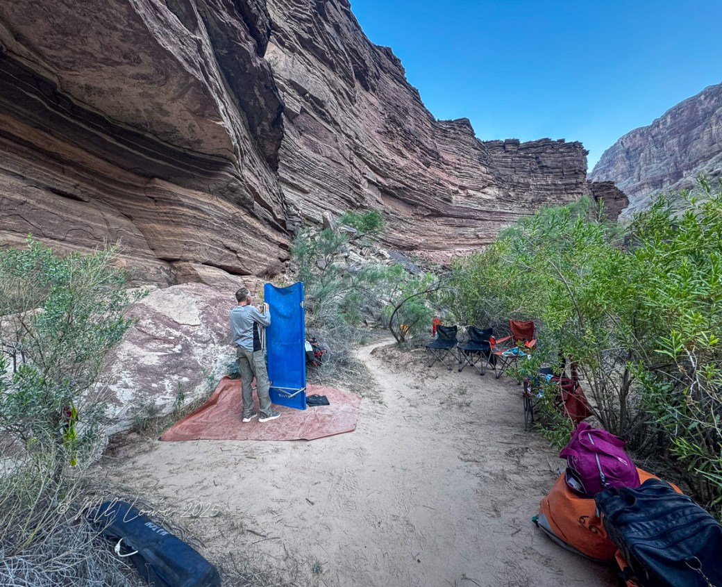 A person using a portable privacy screen in a campsite nestled between rocky canyon walls, with camping chairs and gear visible nearby.
