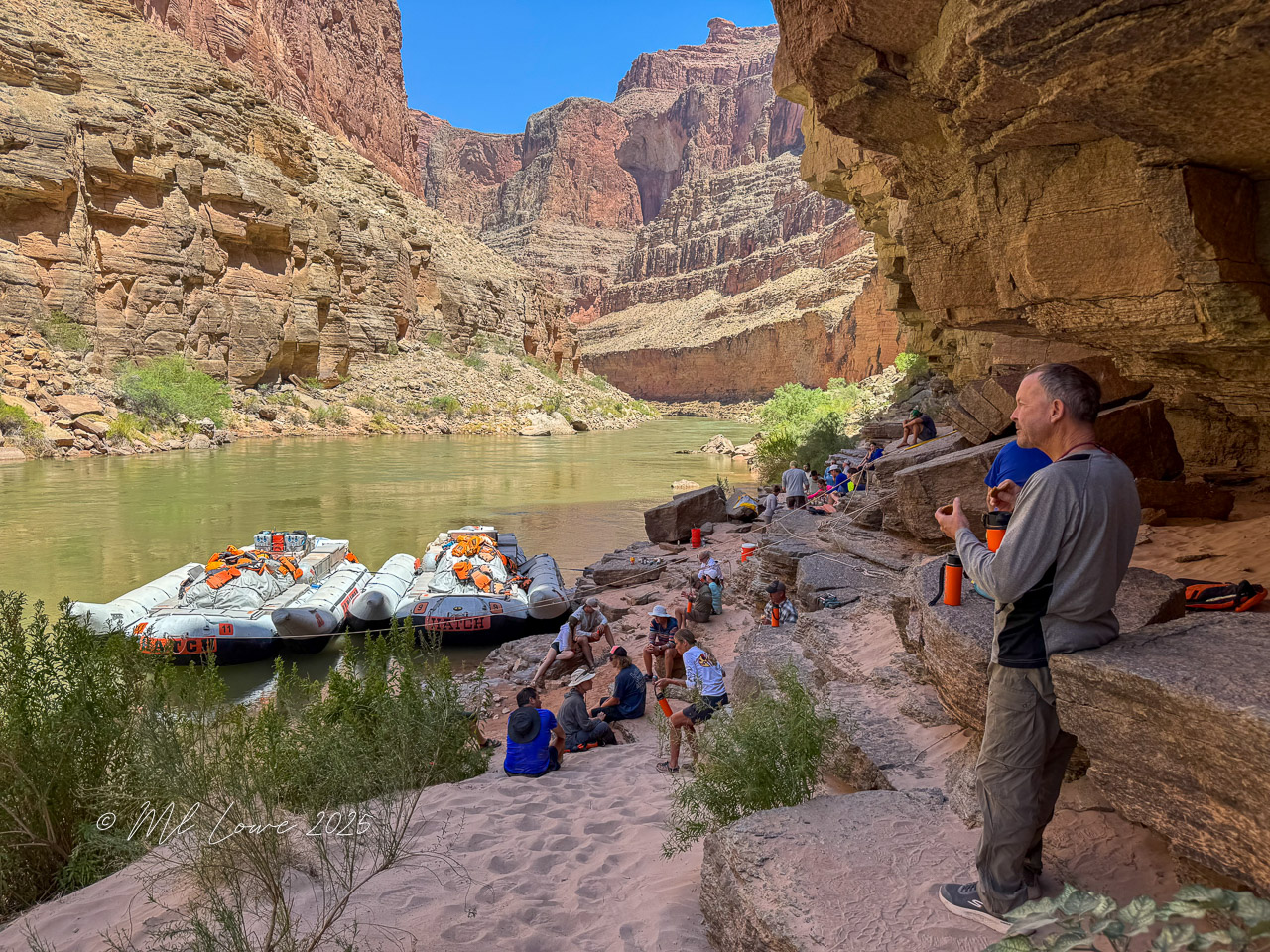 Rafting group relaxing on the sandy bank of the Colorado River, with motorized rafts anchored nearby and steep canyon walls in the background.