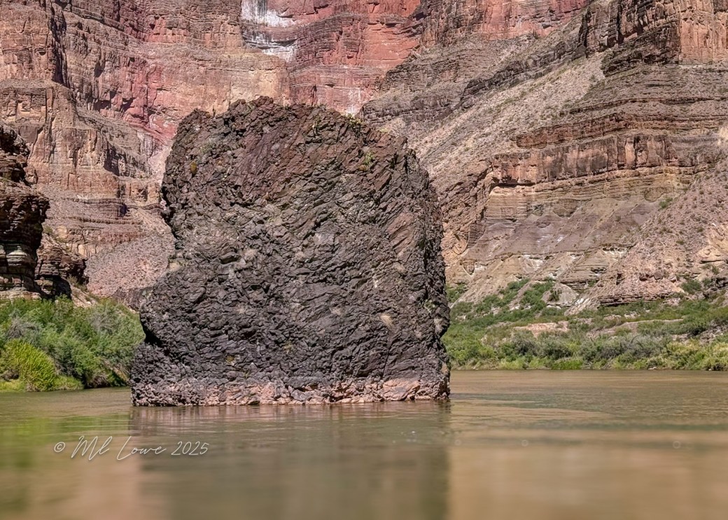 A large rock formation rising from the Colorado River, surrounded by steep canyon walls with layers of colorful rock and vegetation along the water's edge.