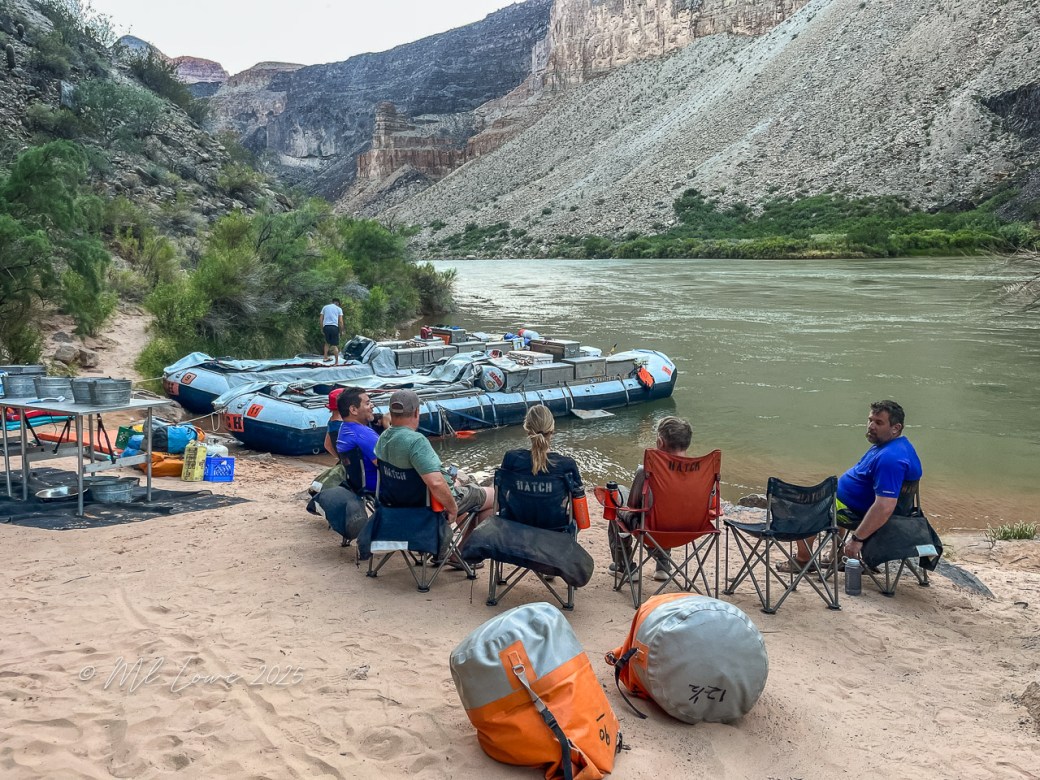 A group of people sitting on beach chairs by the Colorado River, with motorized rafts in the background and steep canyon walls in the distance.