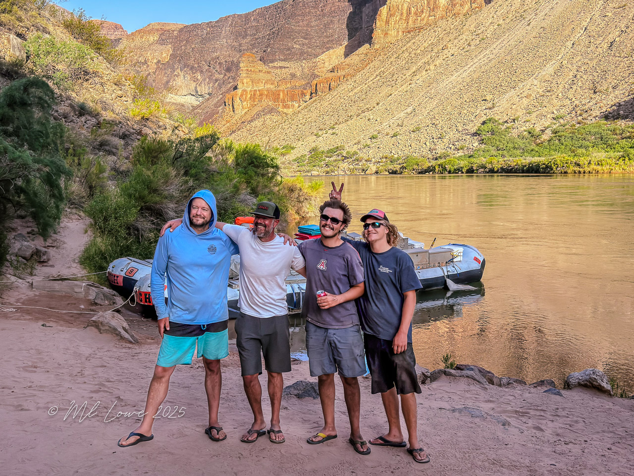 Four men posing together by the Colorado River, with rafts in the background and cliffs towering behind them.