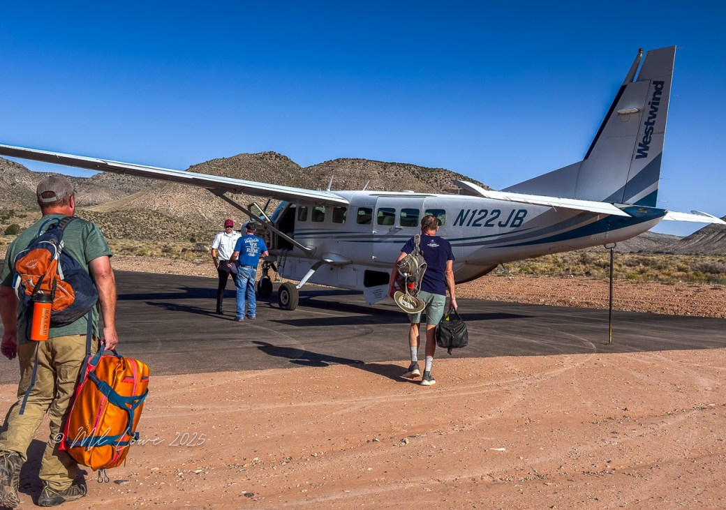 Two men walk towards a small plane parked on a dirt runway in a desert landscape, with mountains in the background.