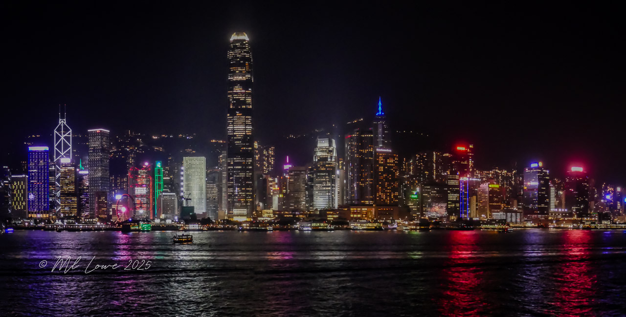 A panoramic view of the Hong Kong skyline at night, showcasing brightly lit skyscrapers and reflections on the water.