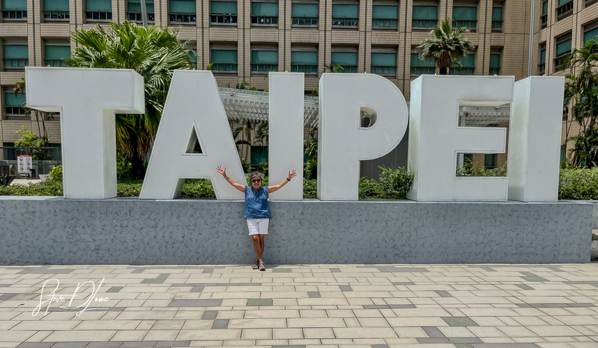 Person posing next to a large sign that reads 'TAIPEI' in a vibrant outdoor setting.