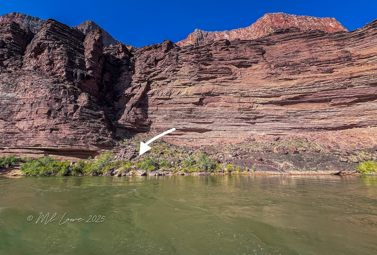 A view of a colorful rock formation alongside a river, with lush greenery at the base and a clear blue sky above.