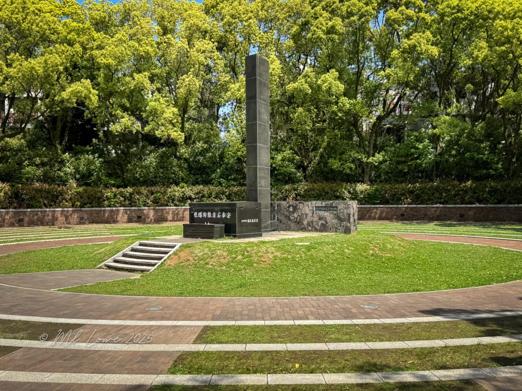 A memorial stone standing tall in a serene park, surrounded by lush green trees, commemorating the victims of the atomic bombing in Nagasaki.