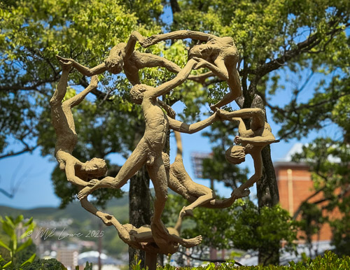 An artistic sculpture depicting human figures engaged in a dynamic circle formation, surrounded by greenery and blue sky.