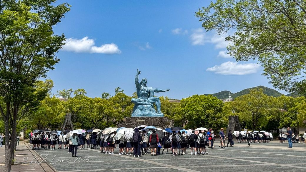 Visitors gathered at the Nagasaki Peace Park, featuring a large blue statue symbolizing peace against a clear sky and surrounded by lush trees.