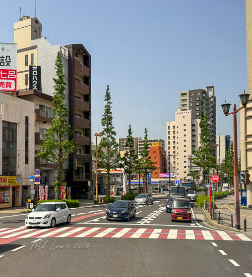 A busy street scene in Japan featuring multiple cars on the road, trees lining the sides, and various buildings in the background. The sky is clear, indicating a sunny day.