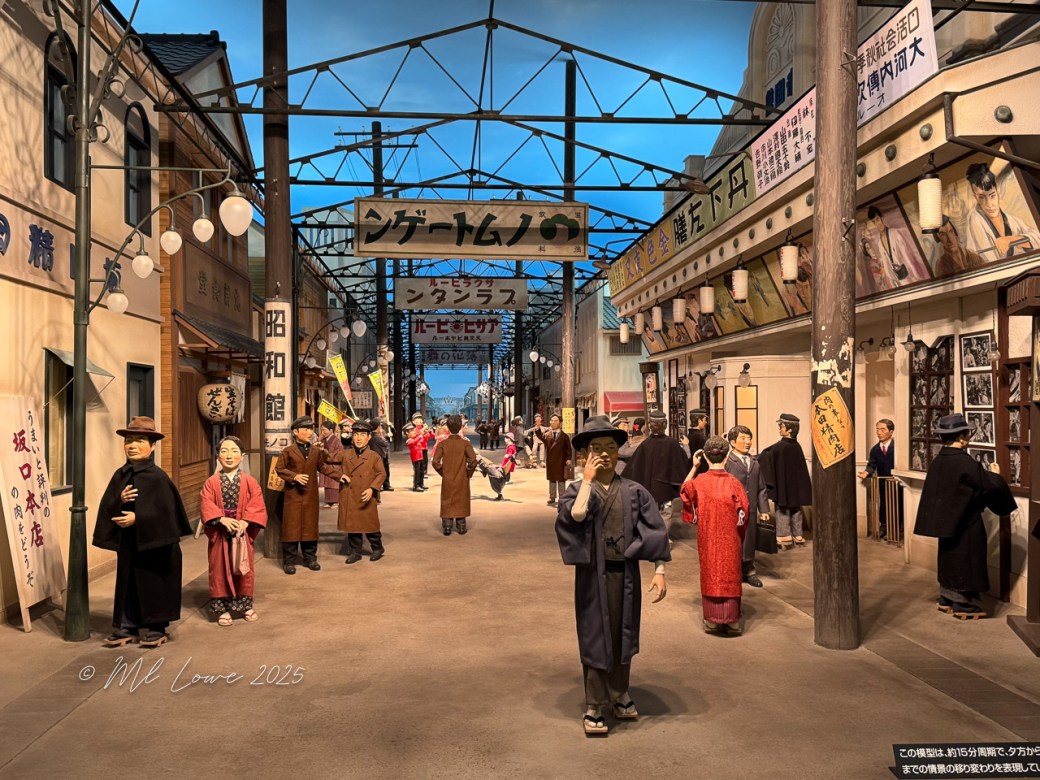A busy street in a historical replica, featuring life-like statues of people in traditional clothing, storefronts, and hanging lights, creating an atmosphere reminiscent of a past era in Japan.