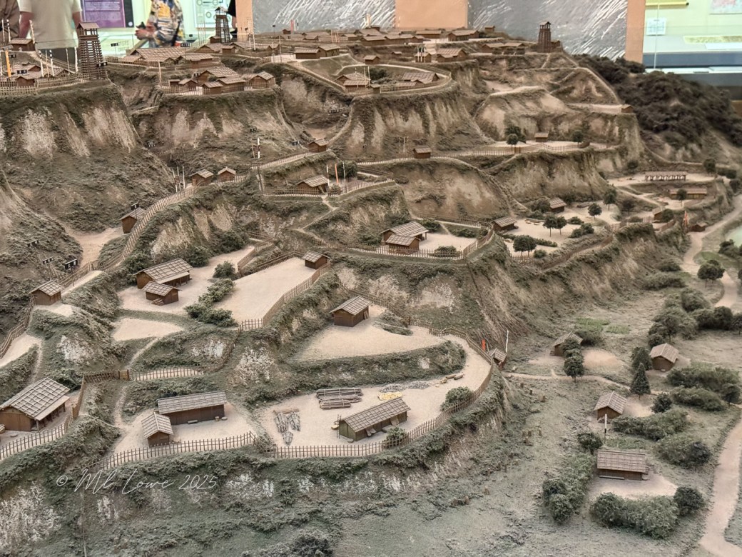 A detailed diorama showcasing an ancient village with various wooden structures built on a hilly landscape, displaying a mixture of trees and dirt paths.