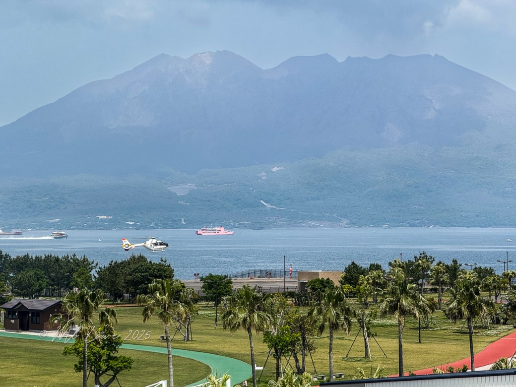 A helicopter flies over a coastal area with a distant view of a volcano and boats on the water, surrounded by green trees and a park.