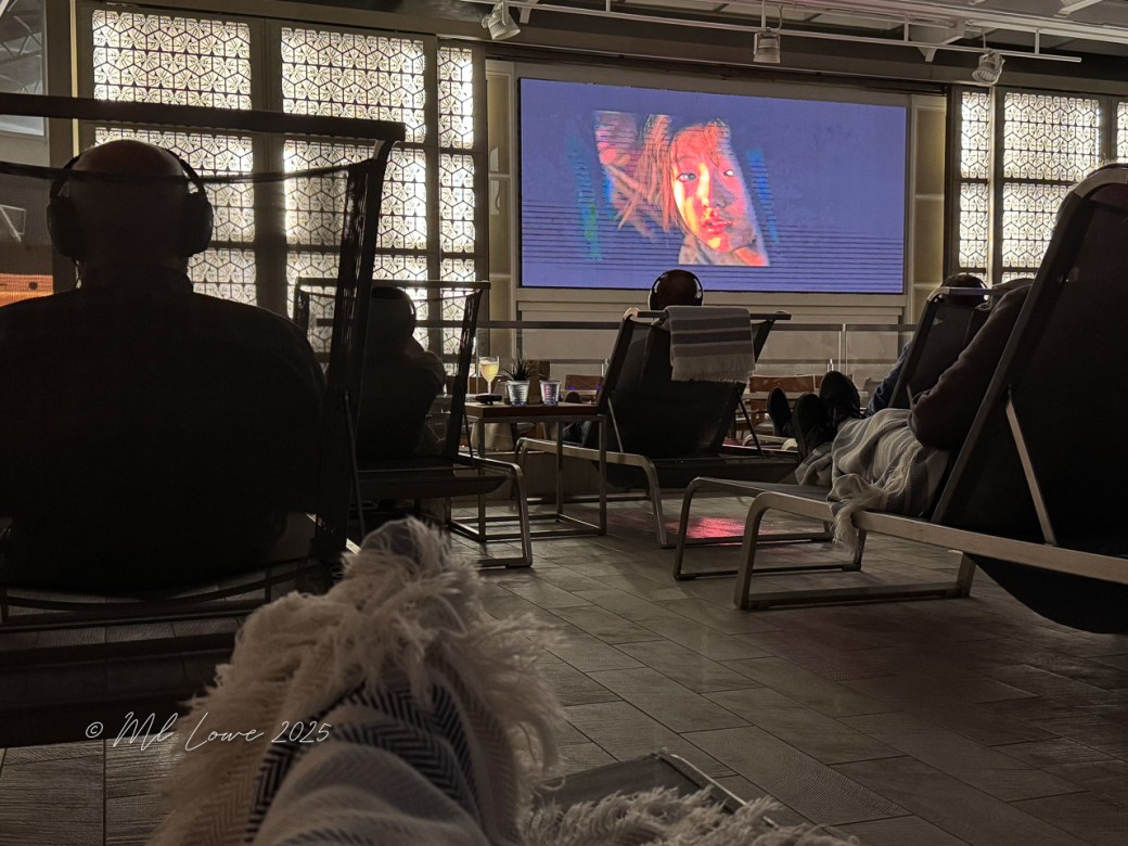 A cozy indoor setting with people relaxing in lounge chairs, watching a movie on a large screen. Headphones are worn by the viewers, and a warm atmosphere is indicated by soft lighting.