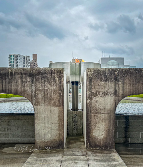 A view of the Eternal Flame at Hiroshima Peace Memorial Park, framed by concrete structures, with modern buildings in the background.