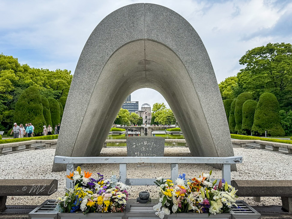 A memorial arch in Peace Memorial Park, Hiroshima, with flowers laid at the base and the Atomic Bomb Dome visible in the background.