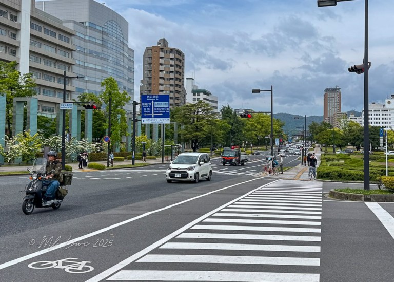 A busy urban street in Hiroshima, showing traffic, pedestrians, and cyclists. Modern buildings and green trees line the road, with traffic signals and road signs visible.