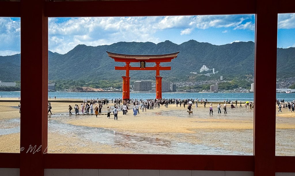A scenic view of the iconic Torii Gate at Itsukushima Shrine on Miyajima Island, framed by red structural elements, with a crowd of visitors exploring the sandy shore and ocean under a bright sky with mountains in the background.
