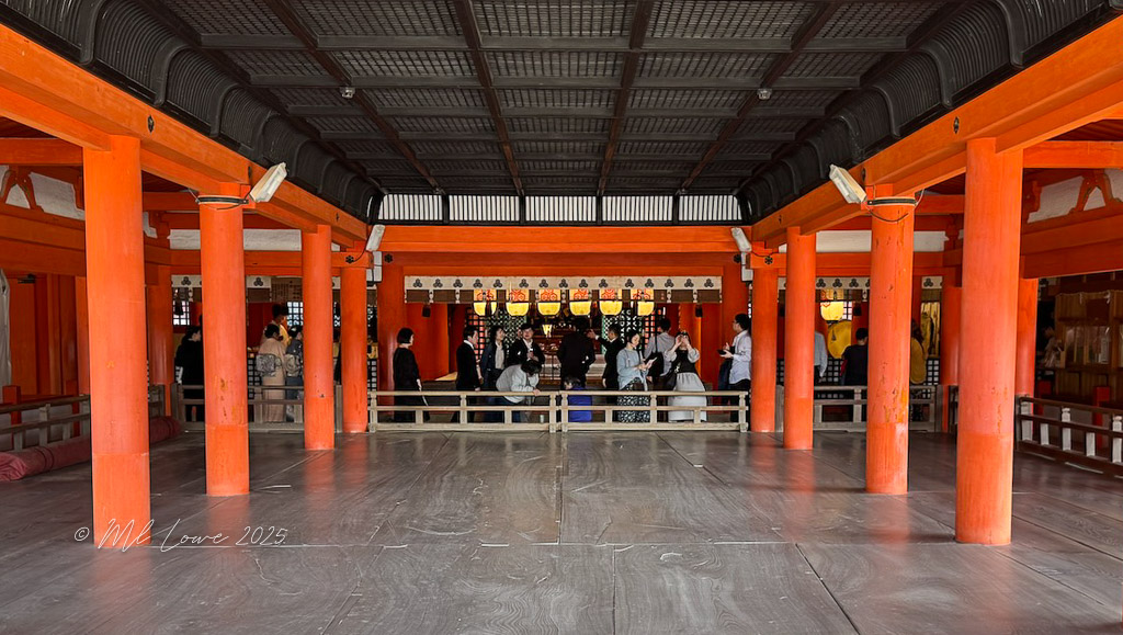 Interior view of a brightly colored Shinto shrine, featuring orange wooden pillars and visitors engaging in activities inside.
