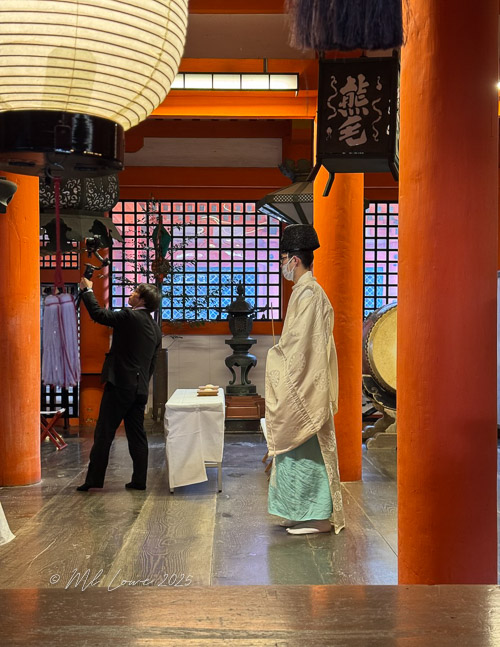 An indoor setting featuring a traditional Japanese shrine with orange pillars and lanterns. In the foreground, a man in a black suit is adjusting a bell or lantern, while a figure in traditional attire, possibly a priest, stands quietly nearby.